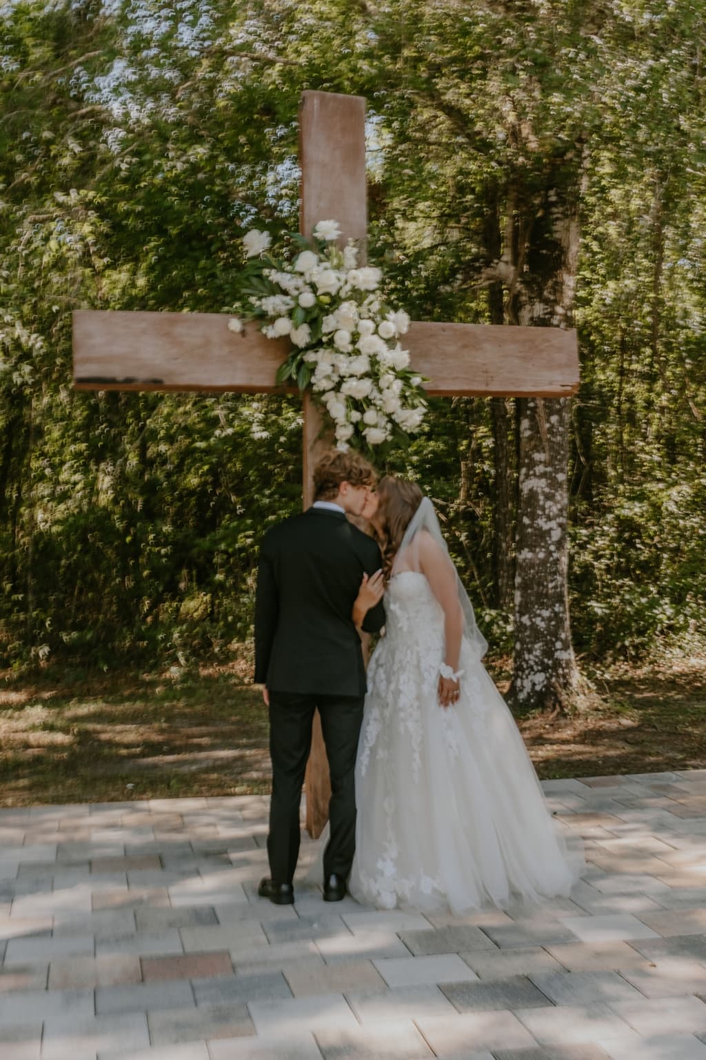 Newlyweds kissing beneath a flower-decorated wooden cross after their ceremony