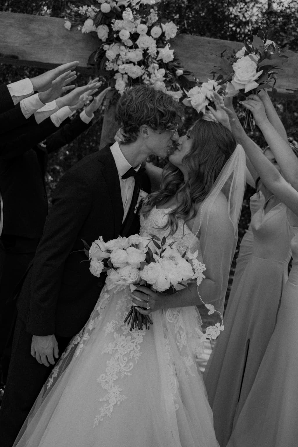 Black-and-white photo of newlyweds kissing beneath raised flowers after the ceremony