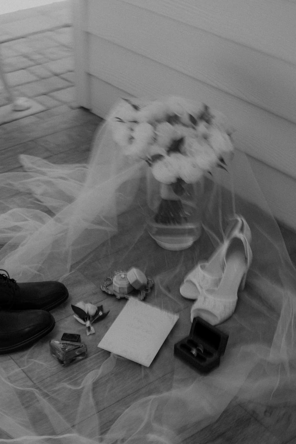 Black-and-white wedding detail flat lay with bouquet, veil, shoes, rings, and invitation